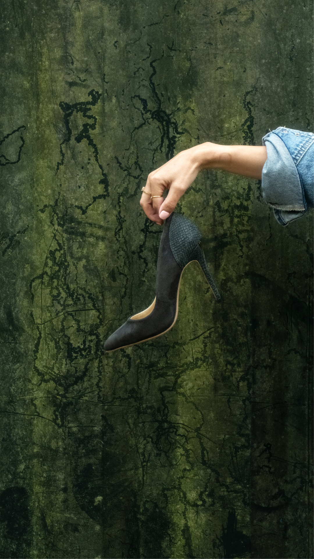 Hand holding a black suede Elandetta pointed heel with textured leather detailing against a deep green backdrop, highlighting colour, silhouette and craftsmanship.