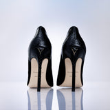 A pair of black high-heeled pumps with a textured finish, shown from the back on a reflective white surface against a light gray gradient background. 