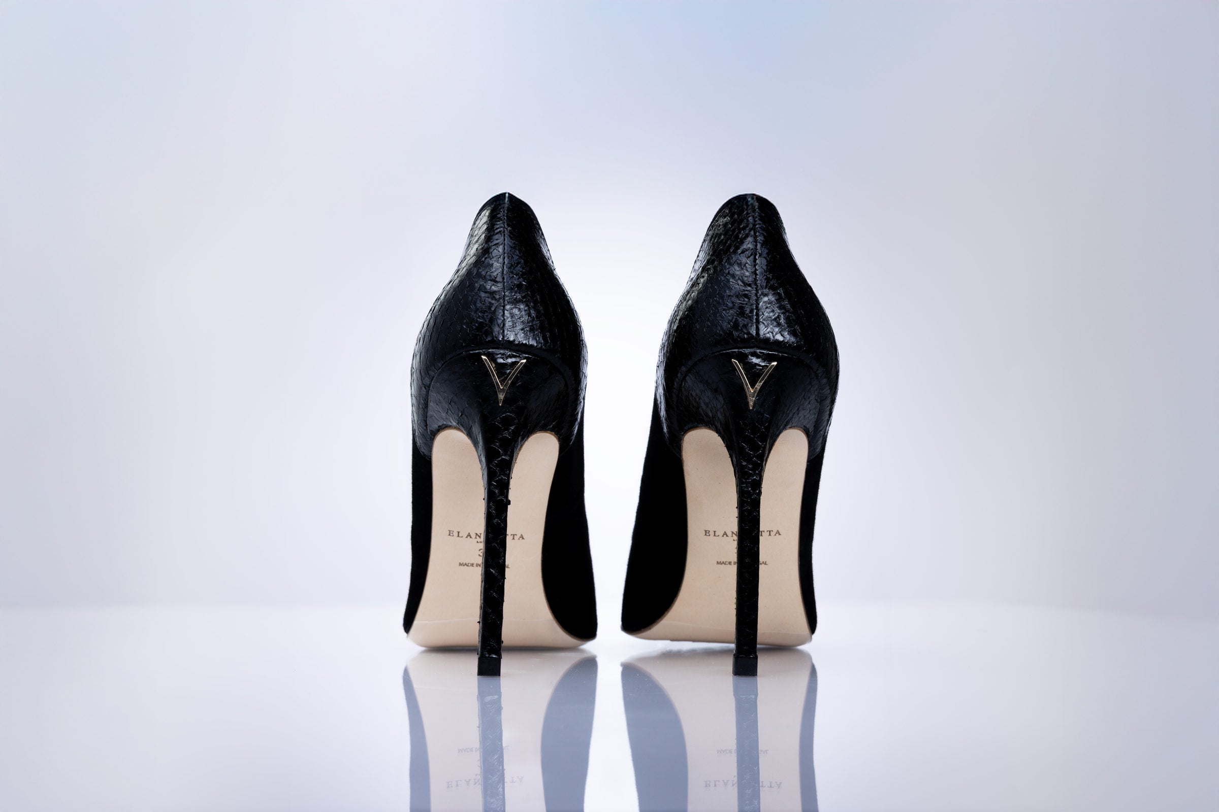 A pair of black high-heeled pumps with a textured finish, shown from the back on a reflective white surface against a light gray gradient background. 