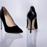 A pair of black high-heeled pumps by Elandetta, shown from the front and back on a reflective white surface. The front shoe features a matte black suede leather, while the back shoe displays a glossy black textured finish with a gold V logo on the heel and a beige insole.
