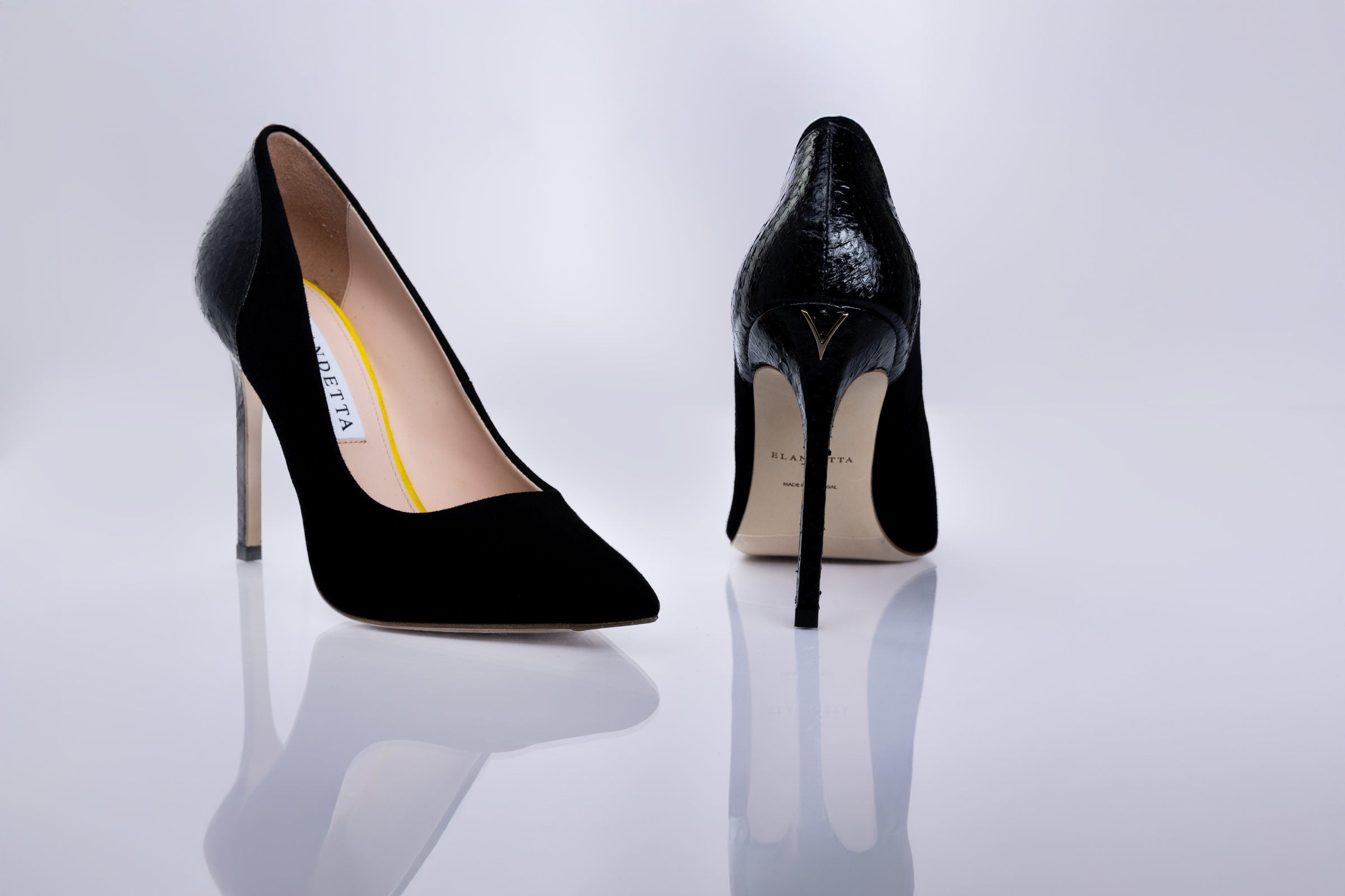 A pair of black high-heeled pumps by Elandetta, shown from the front and back on a reflective white surface. The front shoe features a matte black suede leather, while the back shoe displays a glossy black textured finish with a gold V logo on the heel and a beige insole.