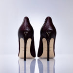 A pair of dark burgundy suede high-heeled pumps shown from the back, featuring a gold-tone logo on the heel counter and leather sole, set against a clean white reflective studio background.