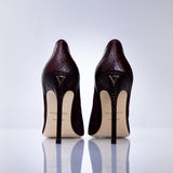 A pair of dark burgundy suede high-heeled pumps shown from the back, featuring a gold-tone logo on the heel counter and leather sole, set against a clean white reflective studio background.