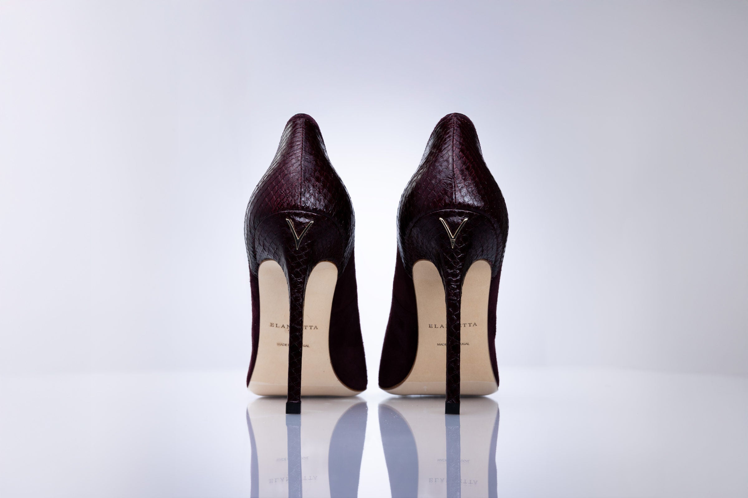 A pair of dark burgundy suede high-heeled pumps shown from the back, featuring a gold-tone logo on the heel counter and leather sole, set against a clean white reflective studio background.