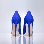 A pair of bright blue high-heeled pumps with a textured snakeskin pattern, shown from the back on a reflective white surface against a light gray background.