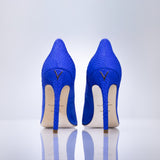 A pair of bright blue high-heeled pumps with a textured snakeskin pattern, shown from the back on a reflective white surface against a light gray background.