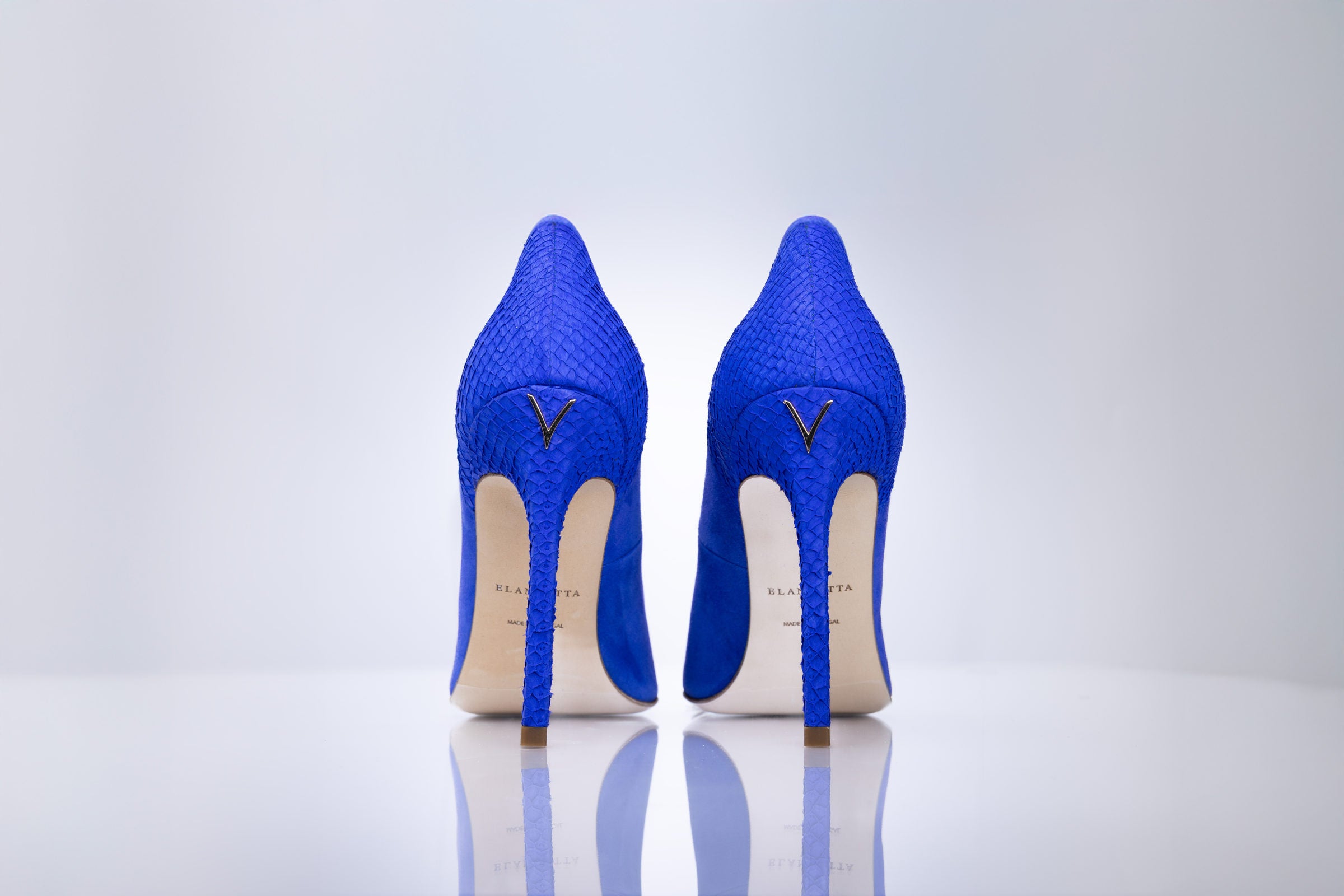 A pair of bright blue high-heeled pumps with a textured snakeskin pattern, shown from the back on a reflective white surface against a light gray background.