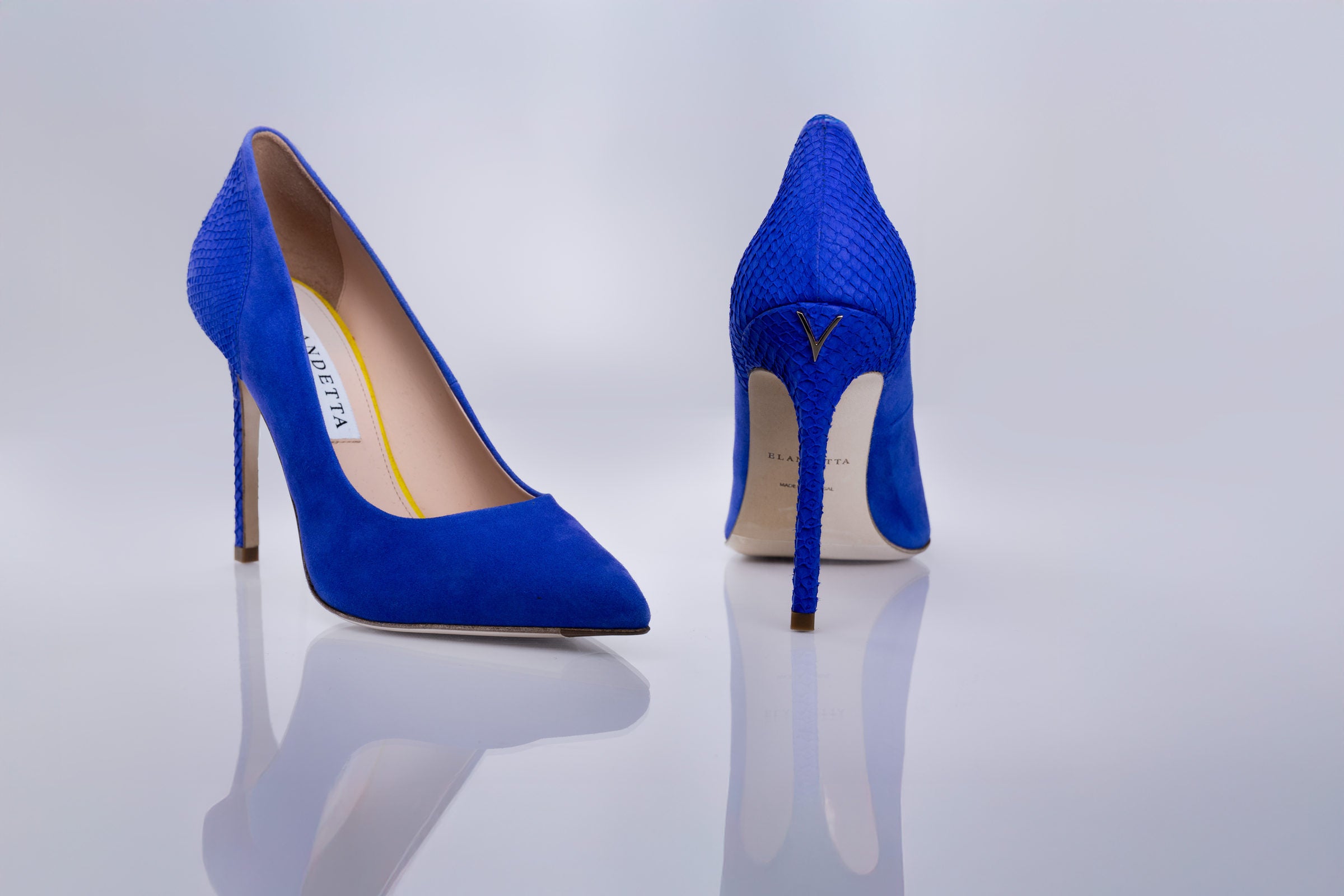 A pair of bright blue pointed-toe high heels with a textured sea leather, shown from the front and back on a reflective light gray studio surface. The brand name logo is visible on the insole.