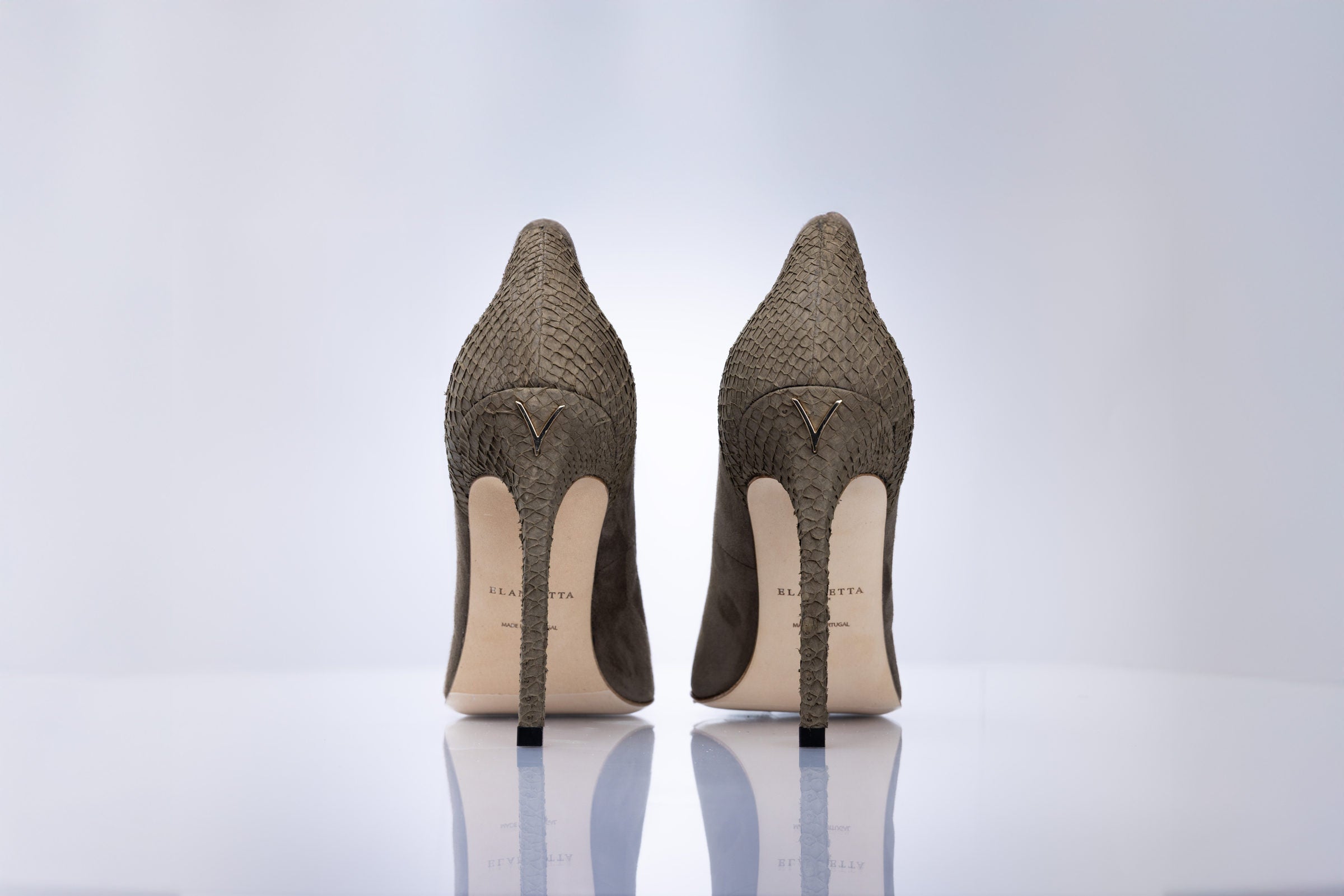 A pair of women's pointed-toe stiletto pumps shown from the back, featuring a taupe suede leather, with a light beige insole and black heels, set against a clean white background.
