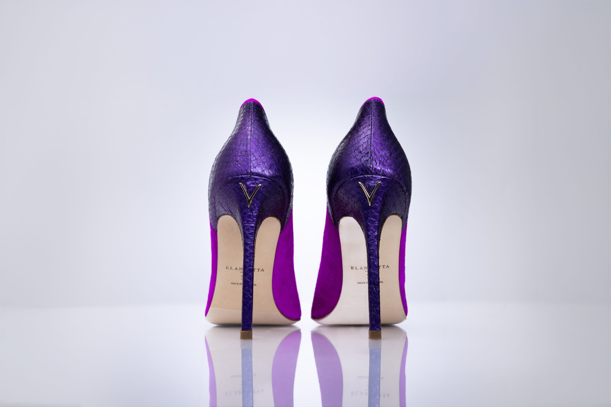 A pair of magenta textured stiletto heels shown from the back on a reflective white surface against a light gray background.