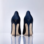 A pair of navy blue high-heeled pumps with a sea leather texture, shown from the back on a reflective white surface against a light gray background. The heels feature a gold-tone V logo on the back of the heel counter and a cream-colored insole.