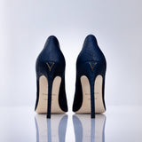 A pair of navy blue high-heeled pumps with a sea leather texture, shown from the back on a reflective white surface against a light gray background. The heels feature a gold-tone V logo on the back of the heel counter and a cream-colored insole.