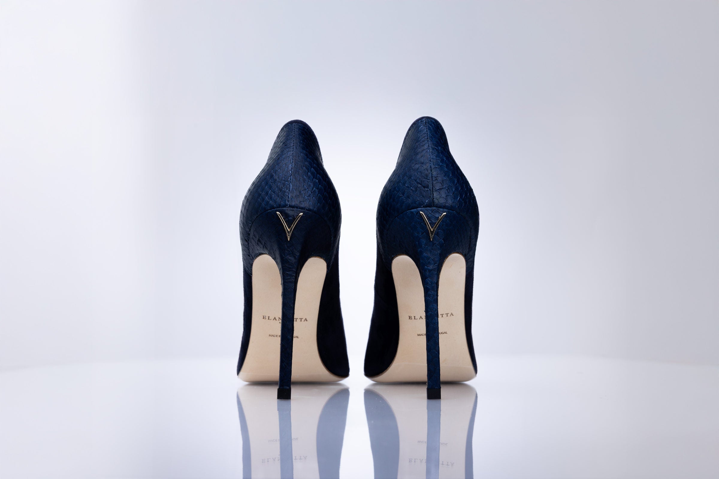 A pair of navy blue high-heeled pumps with a sea leather texture, shown from the back on a reflective white surface against a light gray background. The heels feature a gold-tone V logo on the back of the heel counter and a cream-colored insole.