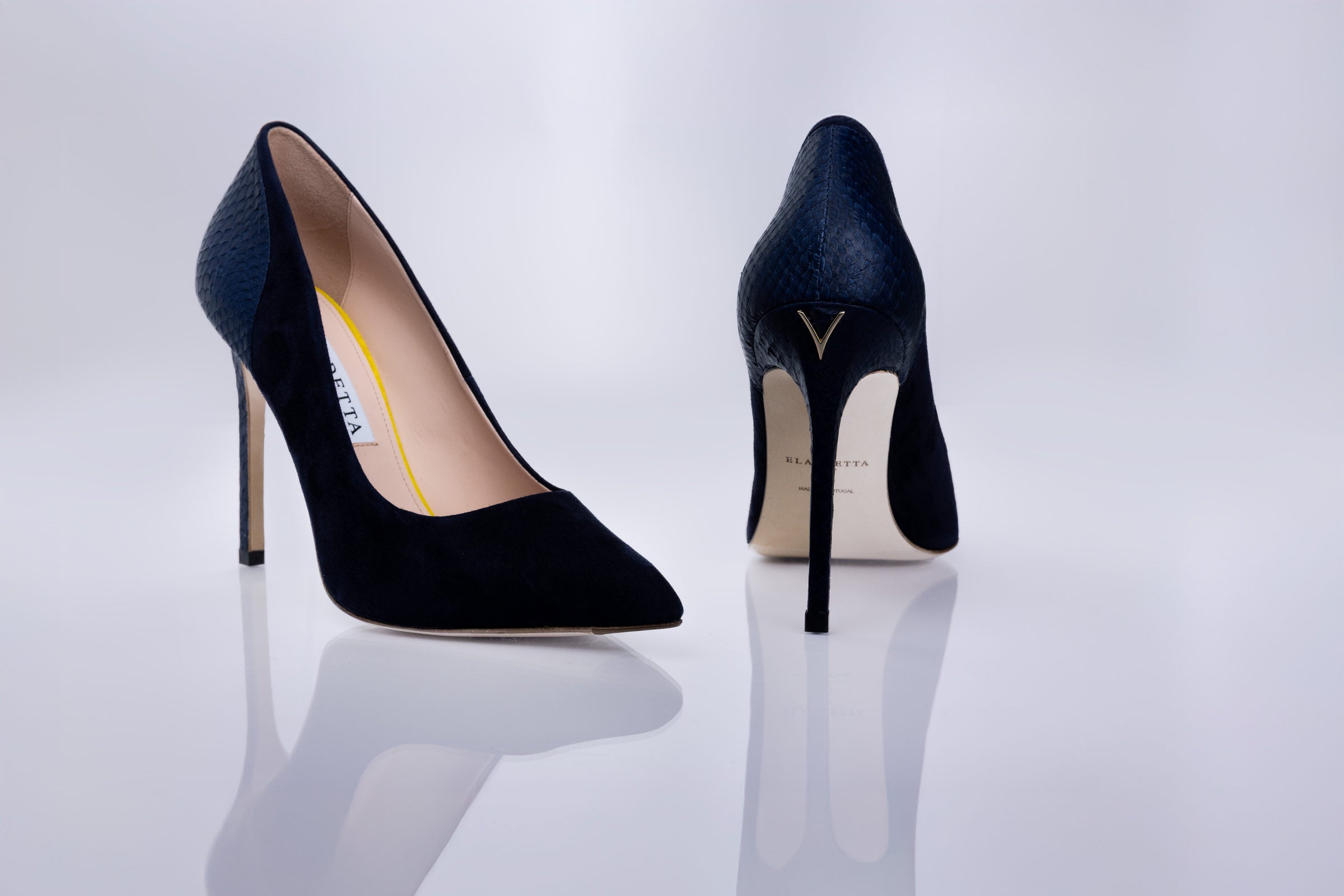 A pair of pointed-toe high heels in dark navy blue textured suede leather, shown from both the side and back angles on a reflective white surface.