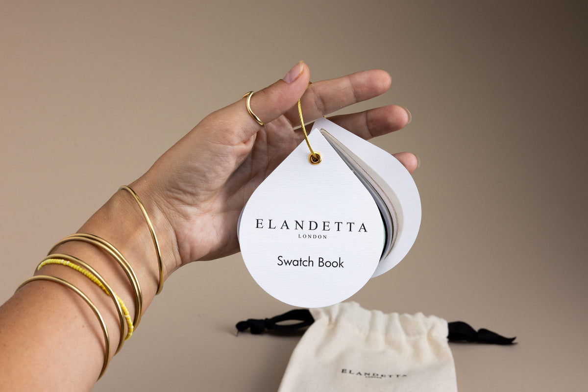 A hand holding an Elandaetta London Swatch Book card, with gold bangles on the wrist and a cream drawstring cotton pouch below on a warm taupe background.