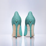 A pair of sage green high heels with a gold V logo on the heels, shown from the back on a reflective white surface against a light gray background.