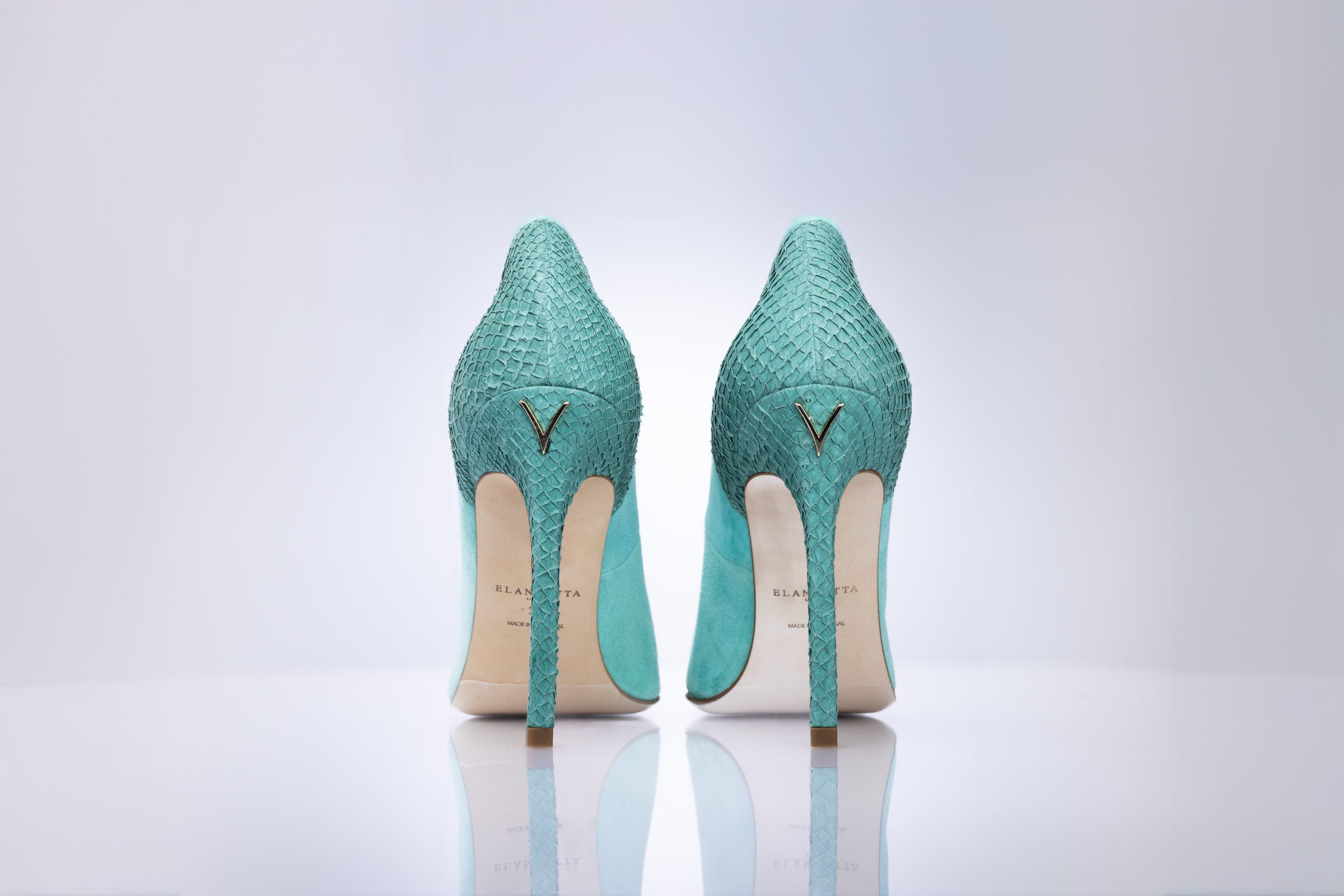 A pair of sage green high heels with a gold V logo on the heels, shown from the back on a reflective white surface against a light gray background.