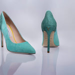 A pair of pointed-toe high heels in a textured turquoise sea leather and suede shown from the side and back on a reflective light gray surface. 