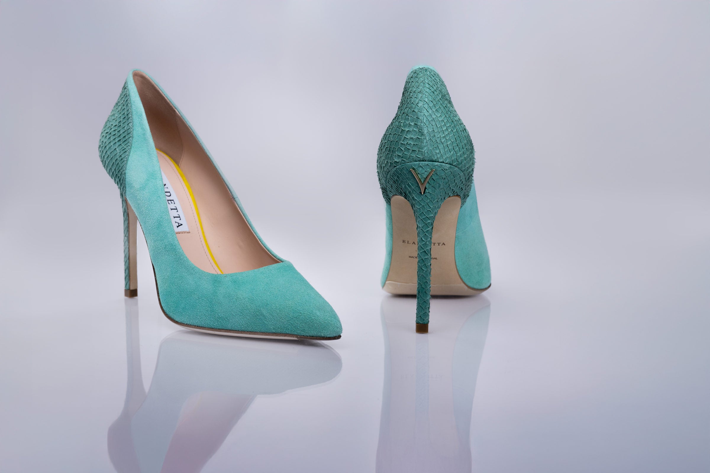 A pair of pointed-toe high heels in a textured turquoise sea leather and suede shown from the side and back on a reflective light gray surface. 