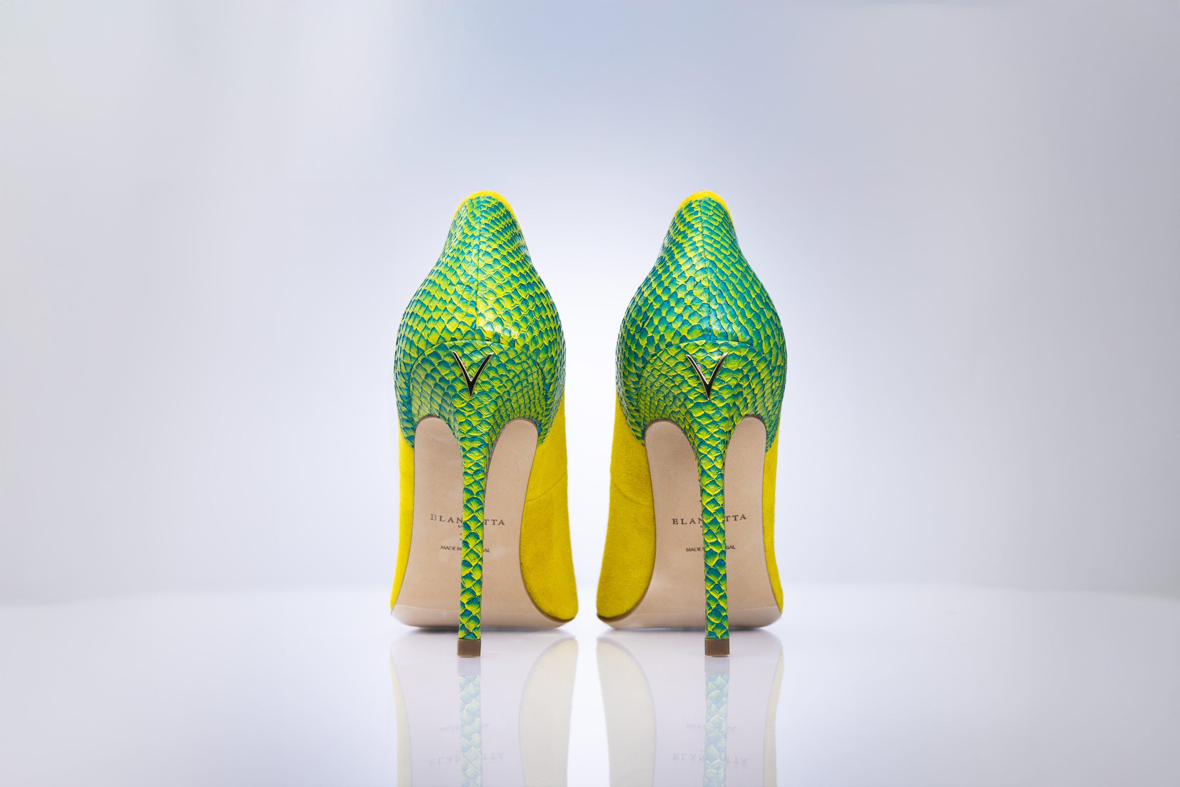 A pair of high-heeled stiletto pumps shown from the back, featuring a vibrant green and yellow python-patterned upper and a bright yellow heel counter, with a beige insole and a small gold ogo on the back.