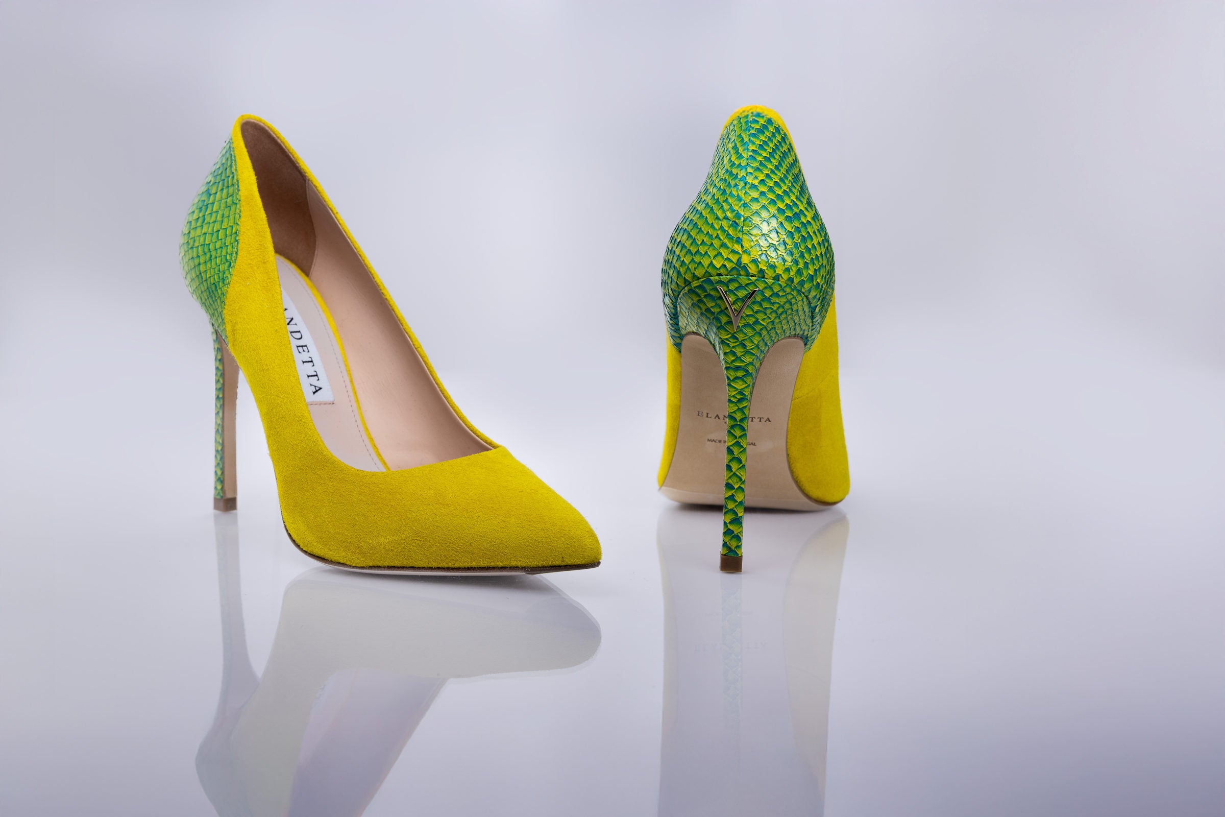 A pair of pointed-toe high heels by Versace, featuring a bright yellow suede-like body and a green snakeskin-patterned upper and heel, displayed on a reflective light gray surface.